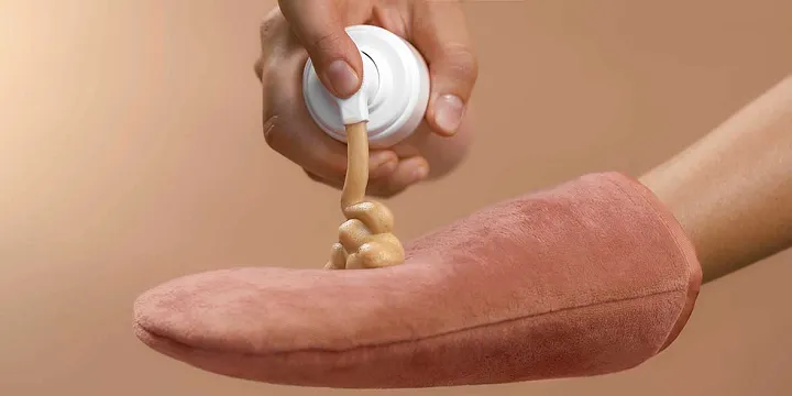 A hand squeezing tanning mousse onto a pink applicator mitt.